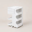 A white three-tier rolling storage cart with drawers on the top left, open shelves on the right, and caster wheels at the base, photographed against a solid light beige background.