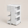 A white rolling storage cart with a top tray, three drawers on the left, three open shelves, and caster wheels, shown on a plain beige studio background.