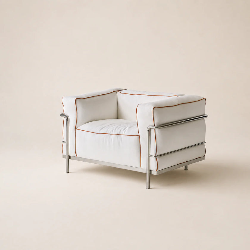 A modern low-profile armchair with white leather upholstery, double chrome steel rails, and thin rust-orange piping, set against a solid warm beige background.