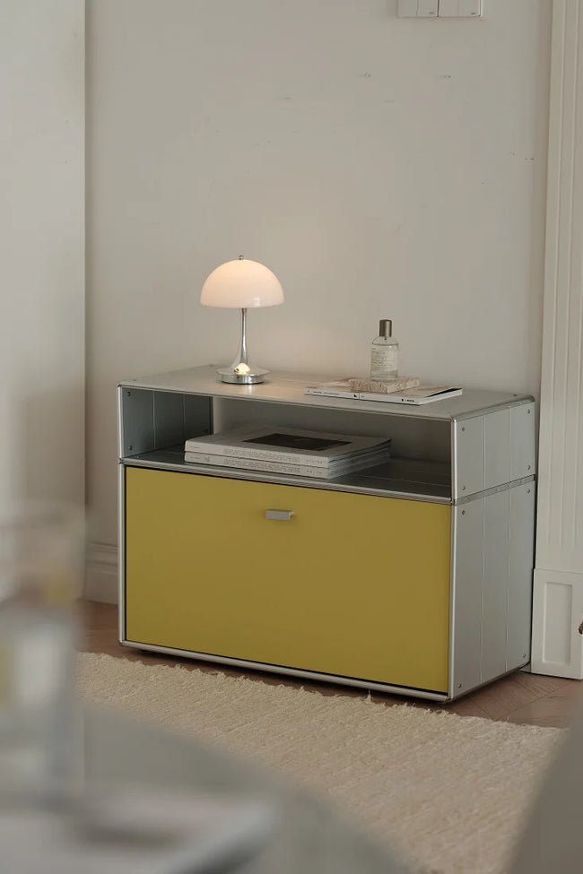 A modern two-tone storage cabinet with a yellow front drawer and a silver metal frame, styled in a minimalist room with a white table lamp and books on top.