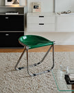 A modern folding stool with a glossy green seat and a chrome metal frame, placed on a textured beige rug in a minimalist room setting.