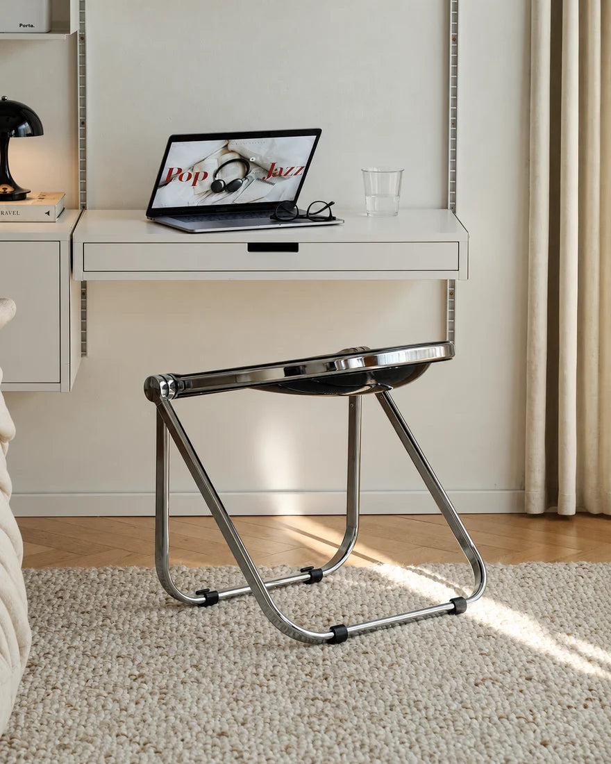 A modern chrome metal folding desk chair with a curved seat, shown in a bright, contemporary room setting with a white desk, laptop, and curtains in the background.