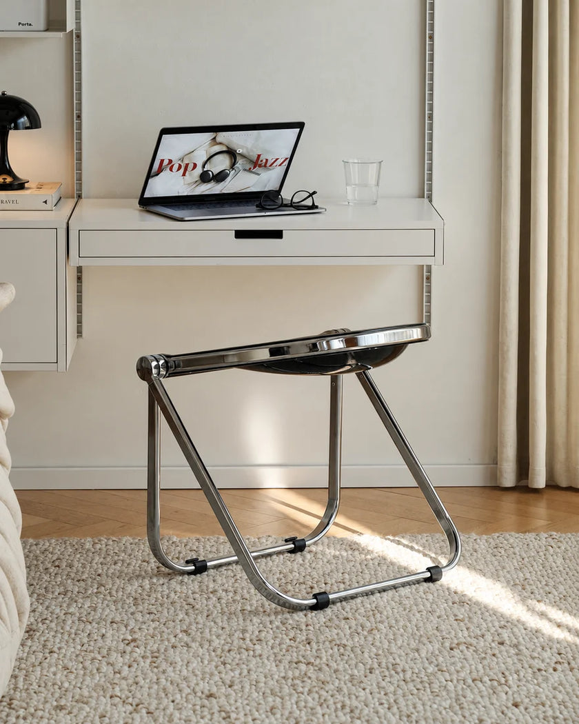 A modern chrome metal folding desk chair with a curved seat, shown in a bright, contemporary room setting with a white desk, laptop, and curtains in the background.