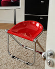 A modern folding chair with a glossy red seat and a chrome metal frame, positioned on a beige carpet in an indoor room setting.