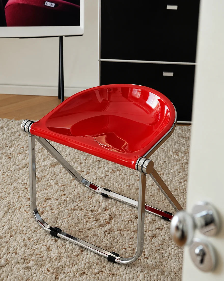 A modern folding chair with a glossy red seat and a chrome metal frame, positioned on a beige carpet in an indoor room setting.