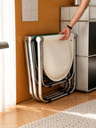 A stack of white folding chairs with chrome metal frames is shown in a home interior setting, with a person's hand reaching to adjust the top chair.