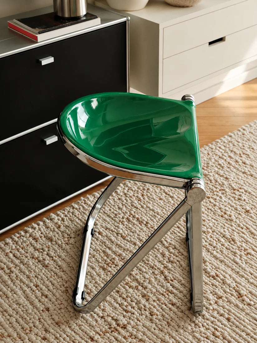 A modern folding chair with a glossy green seat and a chrome metal frame, positioned on a textured beige rug in a contemporary room setting.