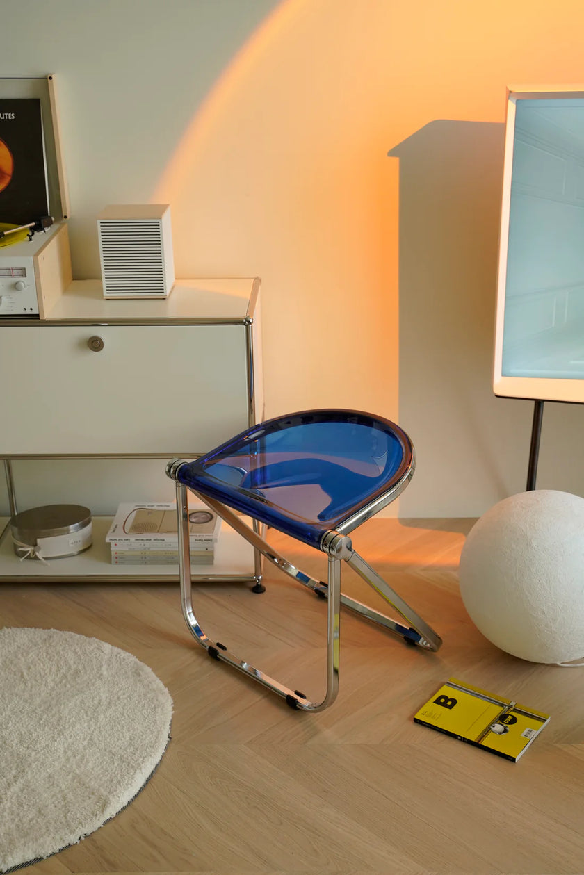 Modern folding stool with blue seat and transparent backrest in a room with a lamp and shelf.