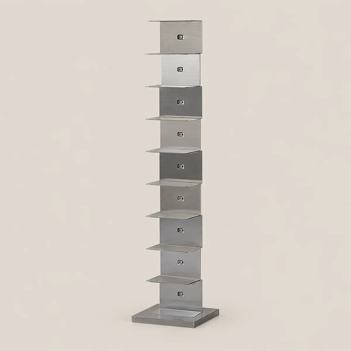 A tall, narrow modern shelf made of brushed silver metal, featuring multiple stacked rectangular compartments, set against a solid light beige background.