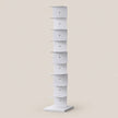 A tall, narrow white display stand with multiple tiered shelves, shown on a plain warm beige background.