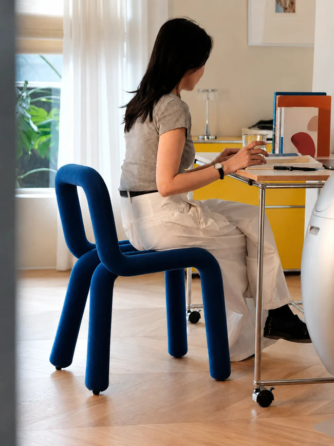 A woman sits on a bold blue upholstered chair at a metal rolling desk in a bright, modern room with light wood floors and a yellow cabinet in the background.