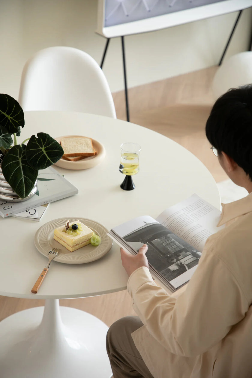Man sitting at a white round modern dining table with a plate of food and a glass of water, reading a magazine.