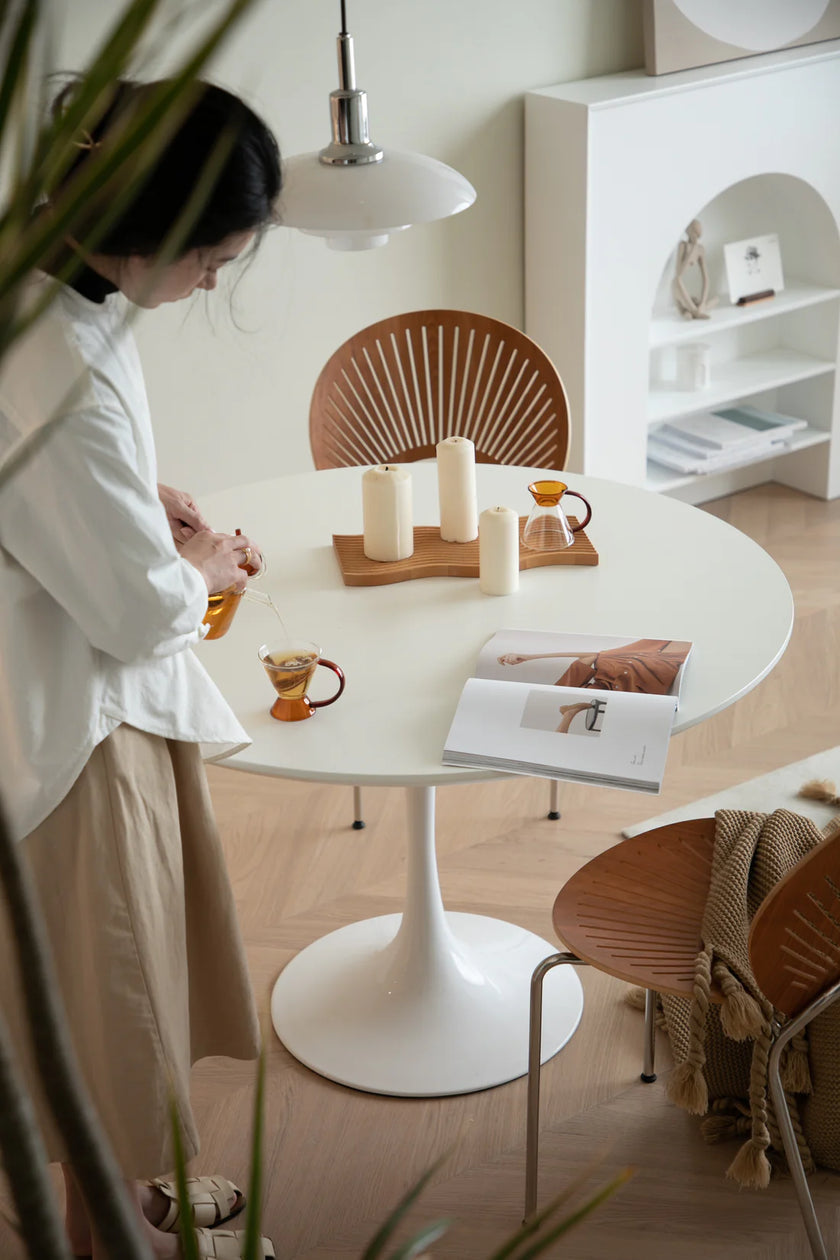 Lady standing near a round white kitchen table with wooden chairs in a modern interior setting.