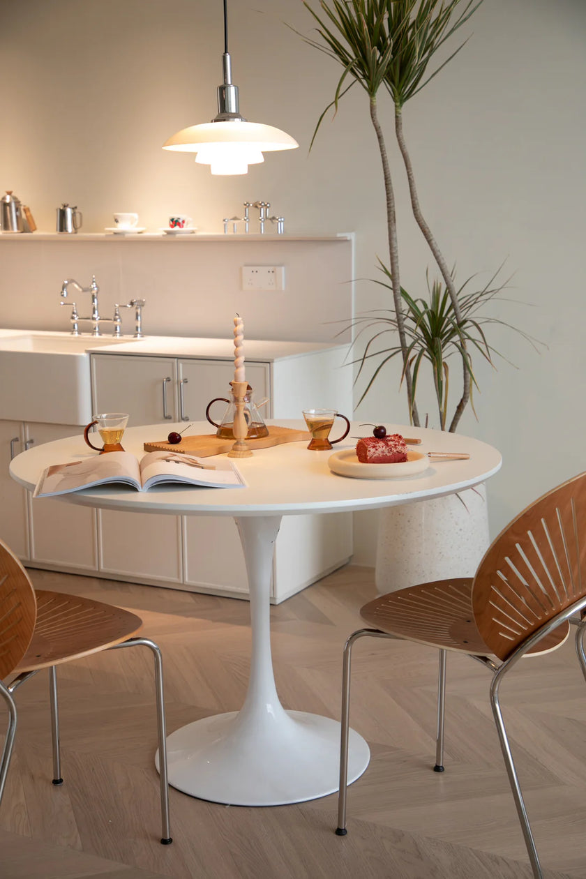 Dining area with a white round kitchen table and wooden chairs, featuring a plant and kitchen in the background.