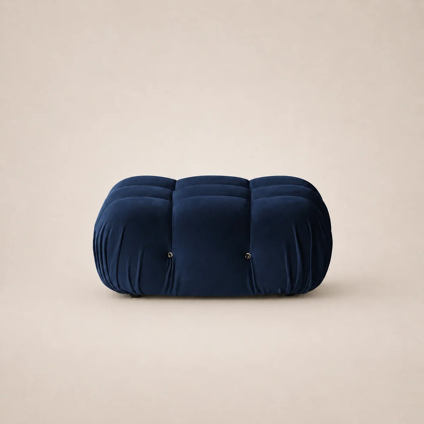 A dark steel blue tufted velvet pouf or ottoman with a rounded rectangular shape, set against a solid beige background.