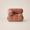 A modern, low-profile modular lounge chair upholstered in muted pink velvet, featuring a rounded, cloud-like silhouette with a separate cylindrical armrest, set against a solid warm beige background.