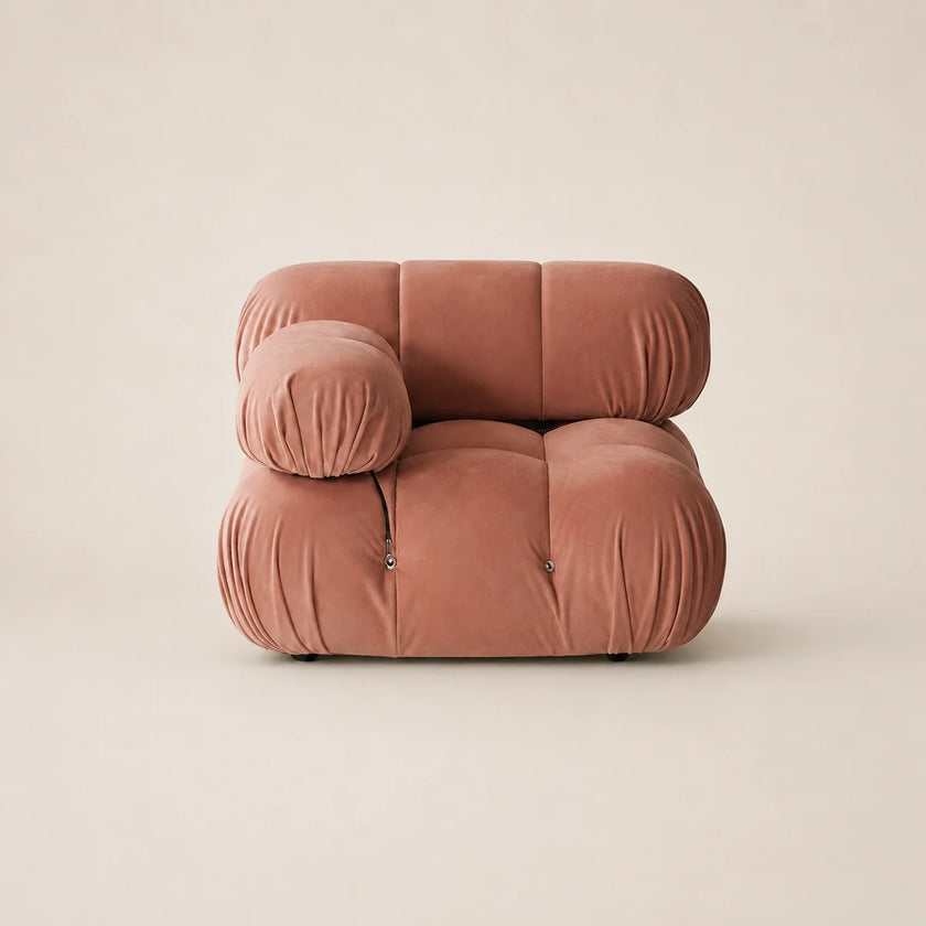 A modern, low-profile modular lounge chair upholstered in muted pink velvet, featuring a rounded, cloud-like silhouette with a separate cylindrical armrest, set against a solid warm beige background.