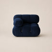 A modern modular floor cushion chair in deep steel blue velvet, featuring a rounded, cloud-like silhouette with zippered pockets, set against a solid warm beige background.