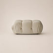 A modern, low-profile cylindrical ottoman or bench upholstered in a soft, tufted fabric in a off white color, set against a solid, warm beige background.