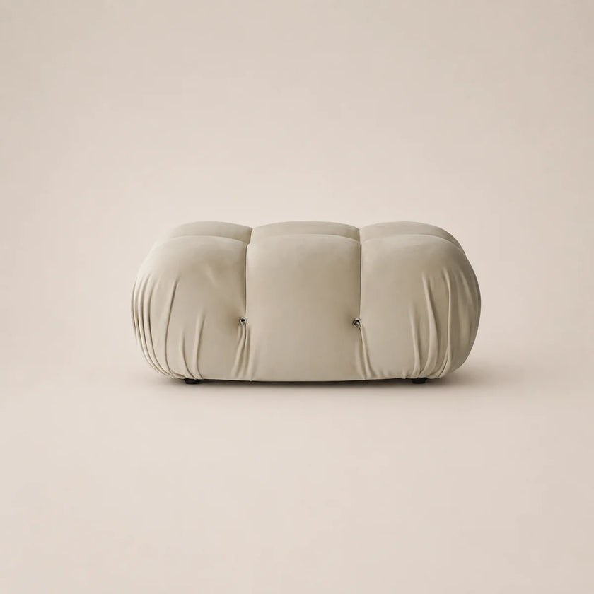 A modern, low-profile cylindrical ottoman or bench upholstered in a soft, tufted fabric in a off white color, set against a solid, warm beige background.