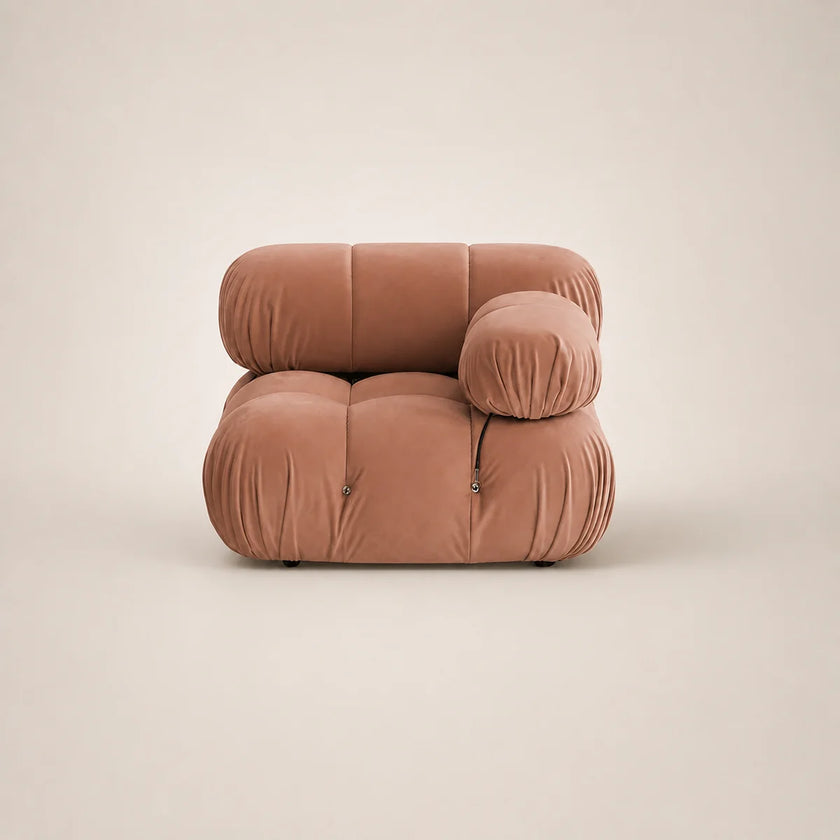 A modern modular lounge chair upholstered in muted pink velvet, featuring a low, rounded blocky silhouette with a separate cylindrical lumbar pillow, set against a solid warm beige background.