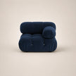 A modern, low-profile modular armchair upholstered in deep steel blue velvet, featuring a rounded, cloud-like silhouette with a separate round back cushion, set against a solid warm beige background.