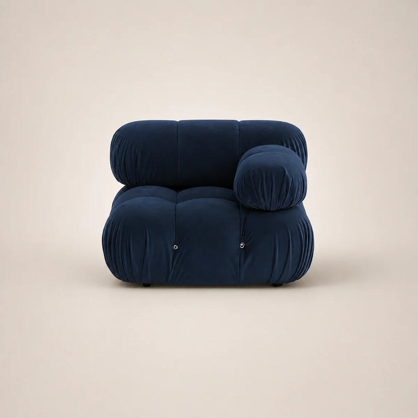 A modern, low-profile modular armchair upholstered in deep steel blue velvet, featuring a rounded, cloud-like silhouette with a separate round back cushion, set against a solid warm beige background.