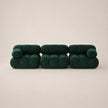 A modern three-seater sofa upholstered in dark forest green velvet, featuring a unique segmented, bubble-like design with cylindrical cushions, set against a solid warm beige background.