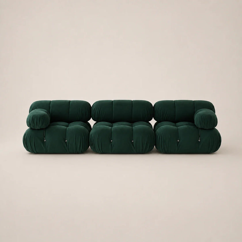 A modern three-seater sofa upholstered in dark forest green velvet, featuring a unique segmented, bubble-like design with cylindrical cushions, set against a solid warm beige background.