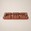 A three-seater modular sofa upholstered in a muted pink fabric, featuring a chunky tufted block design and rounded arms, set against a plain beige studio background.