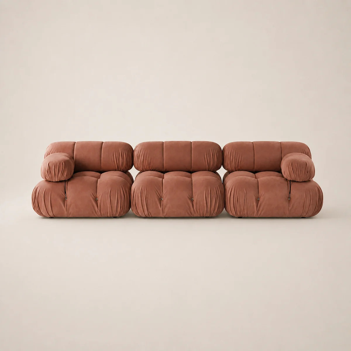 A three-seater modular sofa upholstered in a muted pink fabric, featuring a chunky tufted block design and rounded arms, set against a plain beige studio background.