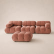 A modern modular sofa set upholstered in muted pink velvet, including a low tufted ottoman in the foreground and three matching cushioned seat and back modules behind it, set against a warm beige studio background.
