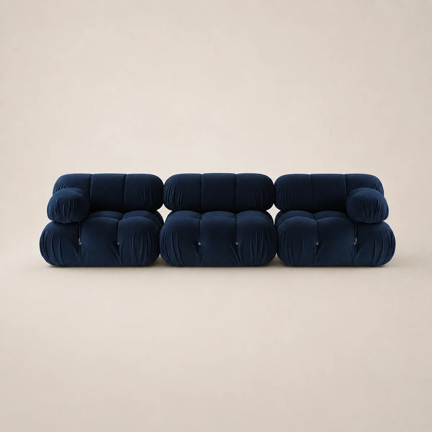 A modern modular sofa upholstered in deep steel blue velvet, composed of three connected tufted cube sections, set against a solid warm beige background.