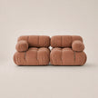 A modern two-seater sofa upholstered in a muted pink velvet fabric, featuring a unique tufted bubble design and small silver metal pull accents on the armrests, set against a solid warm beige background.