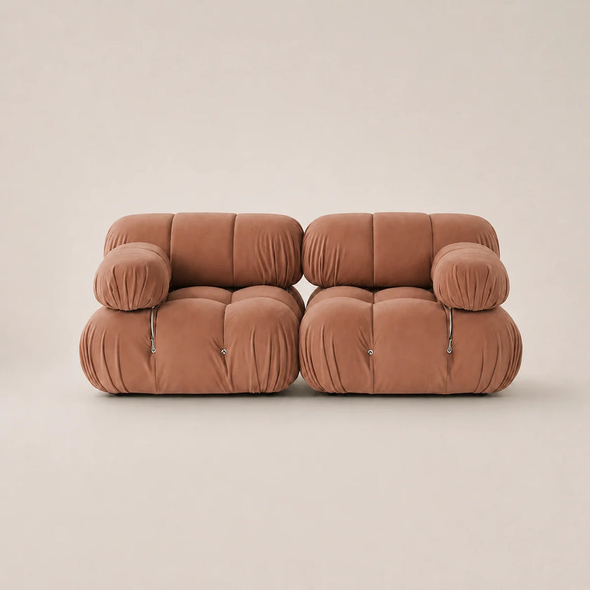 A modern two-seater sofa upholstered in a muted pink velvet fabric, featuring a unique tufted bubble design and small silver metal pull accents on the armrests, set against a solid warm beige background.