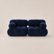 A pair of modern, low-profile modular armchairs upholstered in deep steel blue velvet, set against a solid warm beige background.