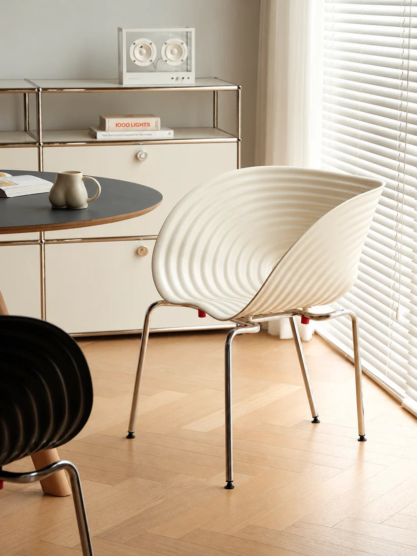 A modern white ribbed shell chair with slim metal legs in a bright, minimalist room with light wood floors, a white cabinet, and a window with blinds.