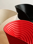 Three modern sculptural bowls with concentric ridges, shown in red, black, and cream on a light wood floor against a neutral wall.