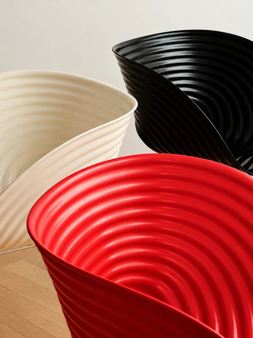 Three modern sculptural bowls with concentric ridges, shown in red, black, and cream on a light wood floor against a neutral wall.