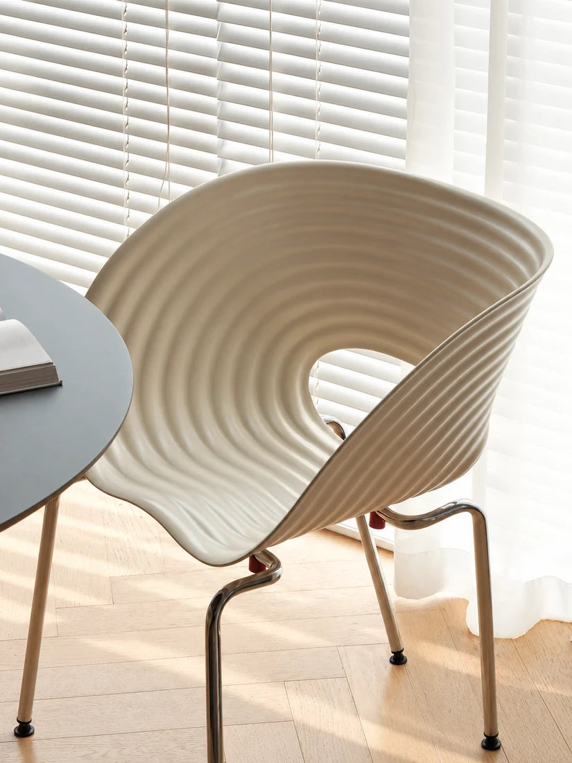 A modern curved accent chair with a ribbed cream-colored shell and slim metallic legs, positioned next to a round gray table in front of white window blinds.