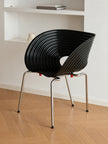 A modern black ribbed shell chair with a chrome metal tubular frame, set on a light wood herringbone floor against a white wall with built-in shelves.