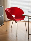 A modern red ribbed plastic chair with chrome legs, positioned in a room with a light wood herringbone floor, white blinds, and a desk.