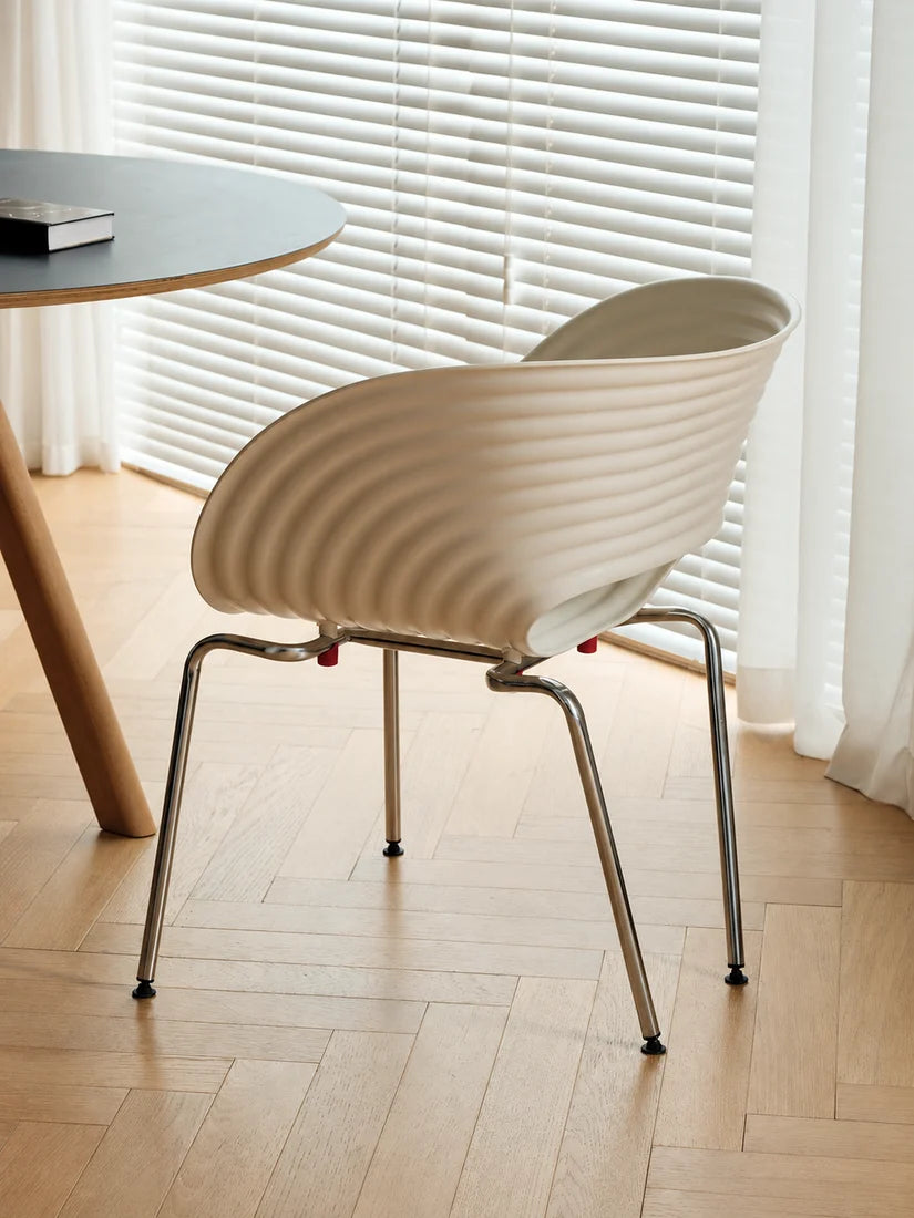 A modern ribbed plastic chair in a warm cream color with a chrome metal frame, shown in a bright room with light wood floors and white window blinds.
