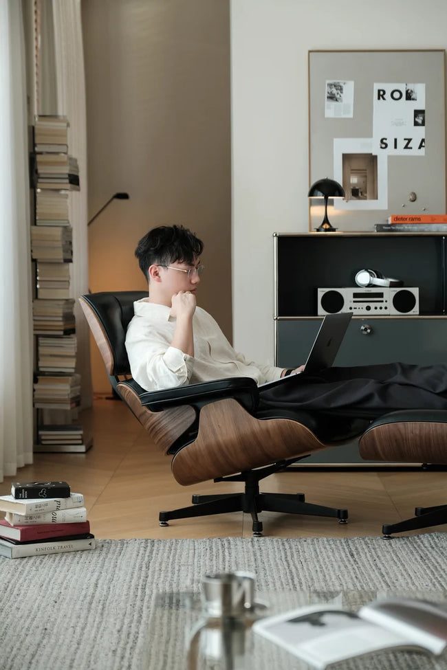 A man wearing glasses and a white shirt relaxes in a brown leather lounge chair with a matching footrest, using a laptop in a modern, stylishly decorated room.