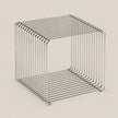 A minimalist, open-frame cube side table made of thin, polished metal wire with a brushed silver finish, set against a solid, warm off-white background.