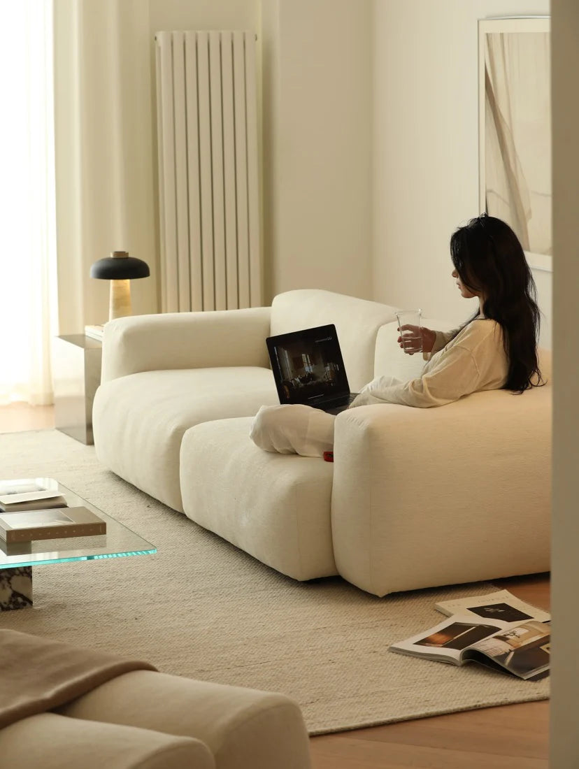 Woman sitting on a white sofa in a modern living room with a laptop and a glass of water.